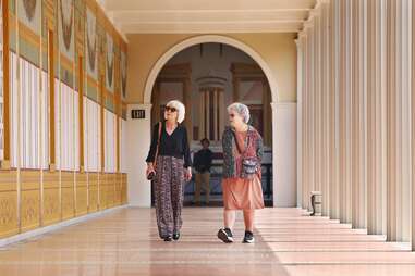 Two visitors walk along a long outdoor corridor decorated with geometric patterns.