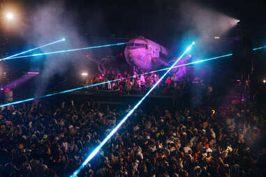 an electronic dance party with laser lights in front of a purple-lit airplane for the after party at splash house