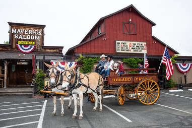 a donkey-drawn carriage in a parking lot in front of the maverick saloon in santa ynez, california, with american flags in the wagon and on the saloon, as in fourth of july decorations